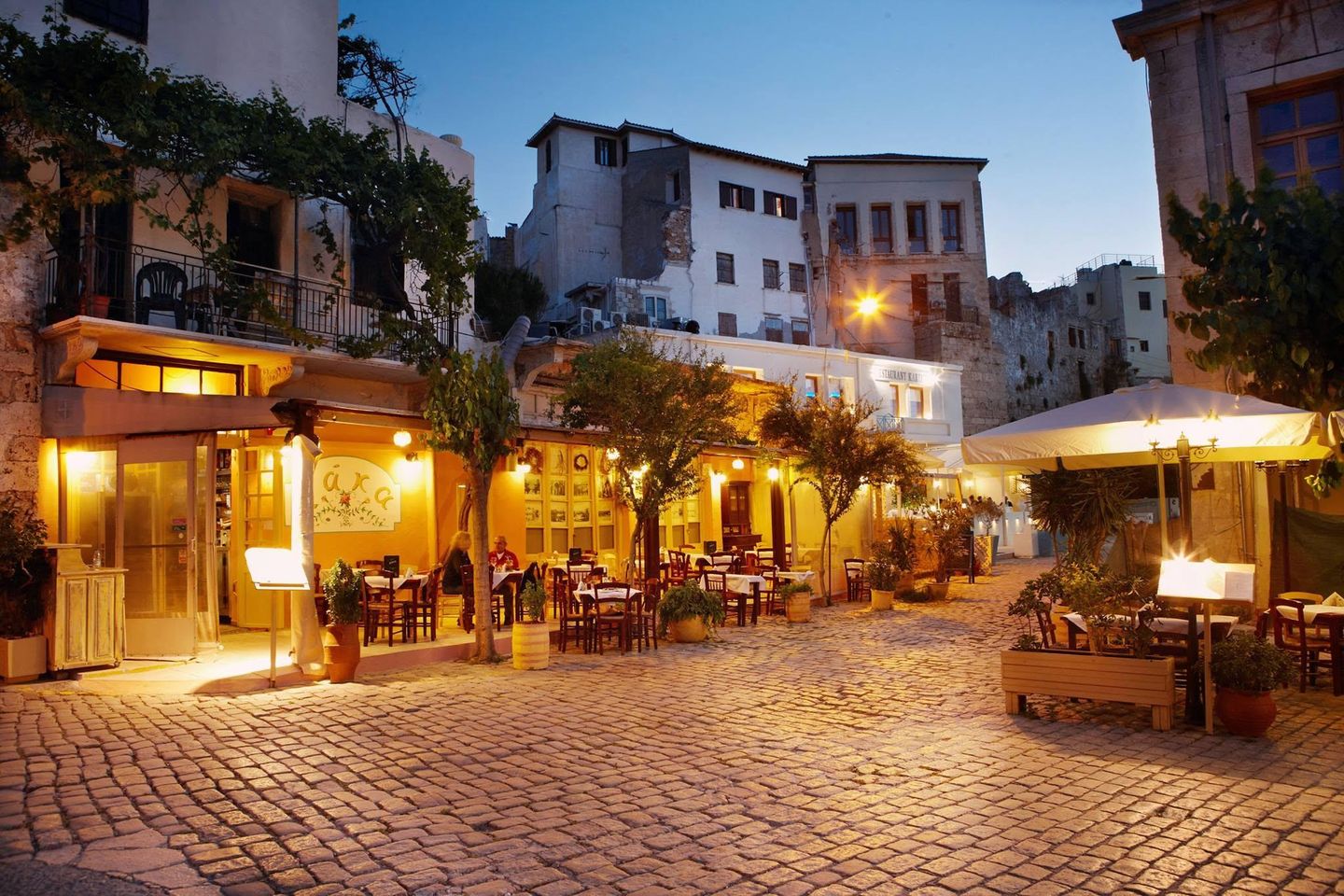 Το στενό τη νύχτα, με φωτισμένες βεράντες και τραπέζια στο καλντερίμι.